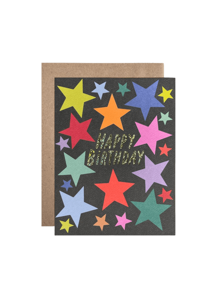 The Happy Birthday Stars Card from Hartland Cards features vibrant, colorful stars on a dark background with glittery gold text, resting in front of a brown kraft envelope | Twentyseven Toronto