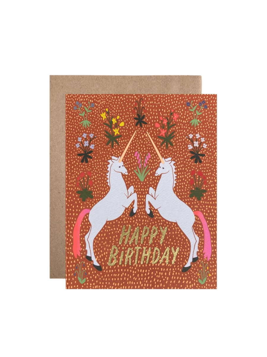 The Happy Birthday Unicorns Card from Hartland Cards features two rearing white unicorns with pink tails facing each other on a textured rust-colored floral background with gold glitter text, paired with a brown kraft envelope | Twentyseven Toronto