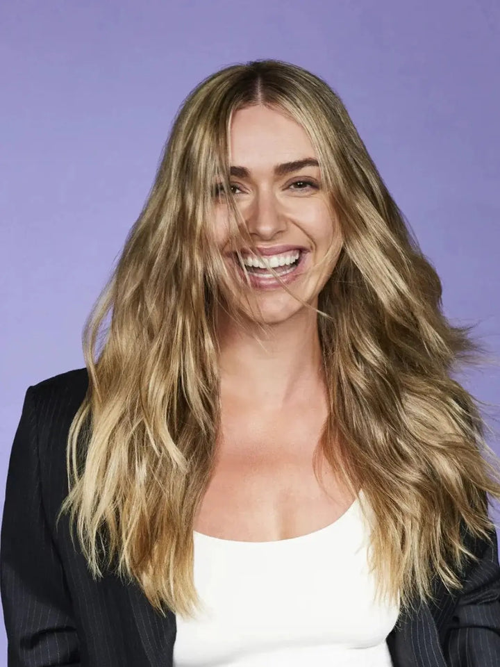A cheerful woman with textured, voluminous blonde hair smiles against a purple backdrop after washing with Hair by Sam McKnight Light Cleanse Hair & Scalp Shampoo | Twentyseven Toronto