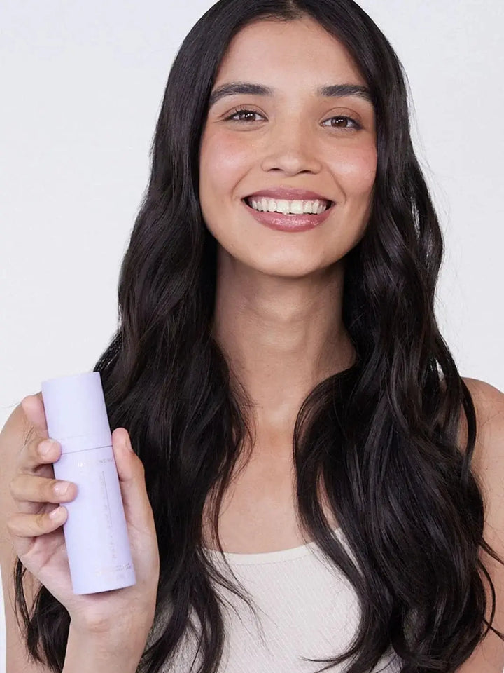 A smiling woman holds a light purple bottle next to her long, wavy dark hair to show the results of using Hair by Sam McKnight Light Cleanse Hair & Scalp Shampoo | Twentyseven Toronto