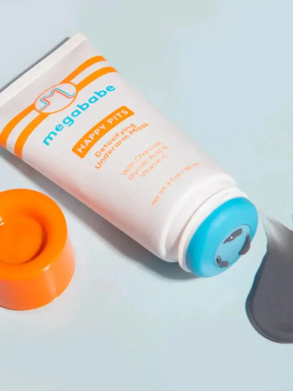 An opened white tube lies flat on a light blue surface next to its orange cap, dispensing a small puddle of grey charcoal formula from the Megababe Happy Pits Underarm Mask | Twentyseven Toronto