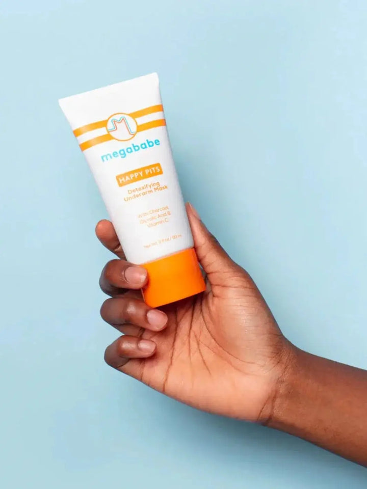 A hand holds up a white tube with an orange cap against a solid light blue background to showcase the packaging of the Megababe Happy Pits Underarm Mask | Twentyseven Toronto