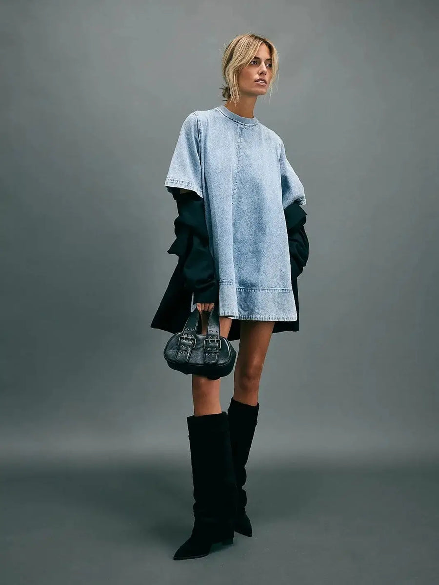 A model showcases the oversized, shift silhouette of the light-wash denim Free People Harlow Mini Dress - Ocean Blue styled with tall black boots and a matching handbag | Twentyseven Toronto