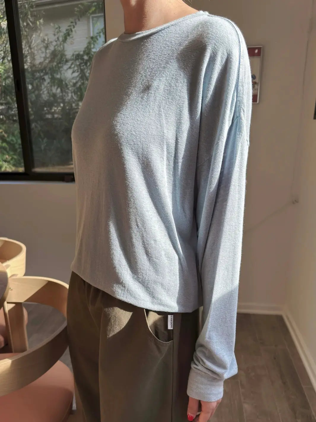 Side view of person wearing a baby blue long-sleeve 'Haze Linen Blend Tee' shirt in 'Moonbeam' and dark green pants from Le Bon Shoppe in an indoor setting | Twentyseven Toronto