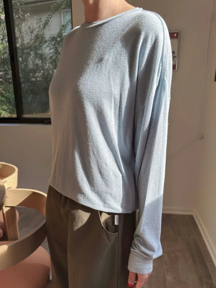 Side view of person wearing a baby blue long-sleeve 'Haze Linen Blend Tee' shirt in 'Moonbeam' and dark green pants from Le Bon Shoppe in an indoor setting | Twentyseven Toronto