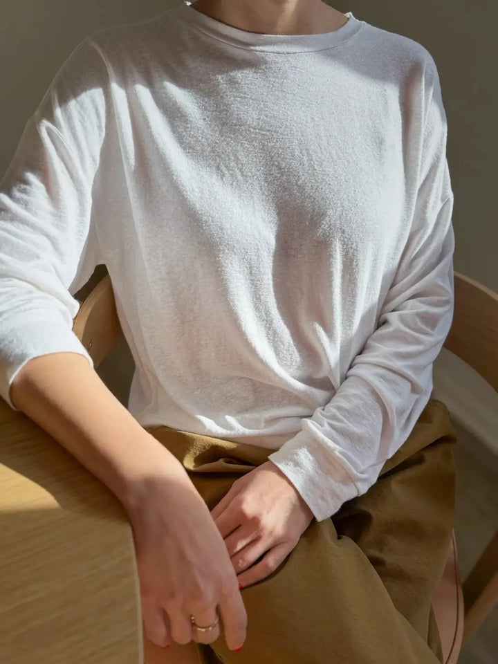 Person wearing a white long-sleeve 'Haze Linen Blend Tee' shirt from Le Bon Shoppe sitting on a wooden chair | Twentyseven Toronto