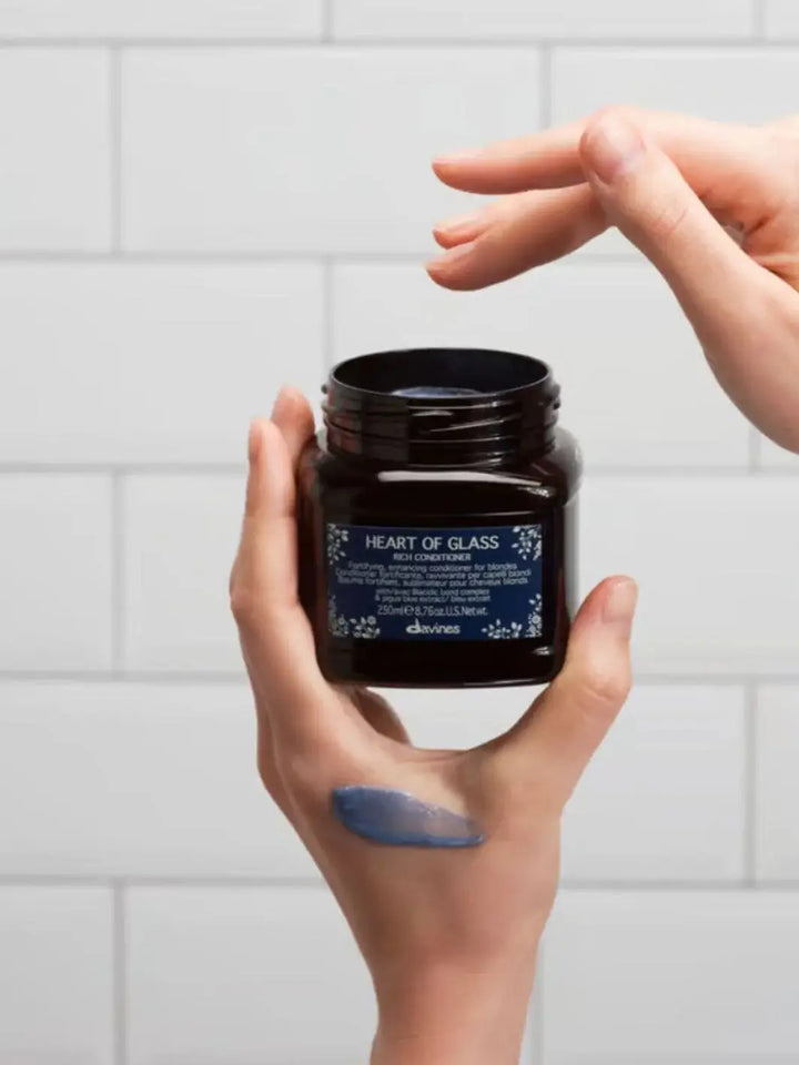 A person holds an open jar of Davines Heart of Glass Rich Conditioner with a smudge of blue cream on their hand against a white tiled background | Twentyseven Toronto