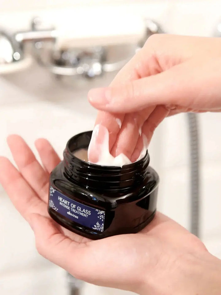 A person dips their fingers into an open dark jar of Davines Heart of Glass Intense Treatment, scooping out thick white hair cream in a bathroom | Twentyseven Toronto