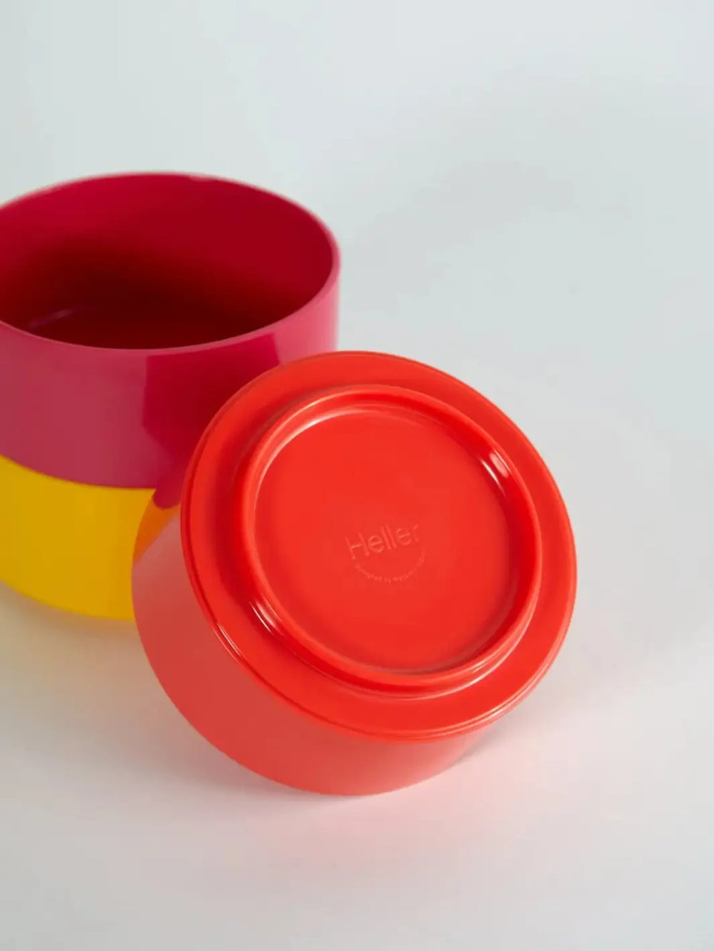 Close-up view showcases the bottom of an orange bowl from the Heller Max Dinnerware Maxbowl in Rainbow - Set of 6, revealing the embossed Heller brand name, alongside stacked yellow and magenta bowls | Twentyseven Toronto