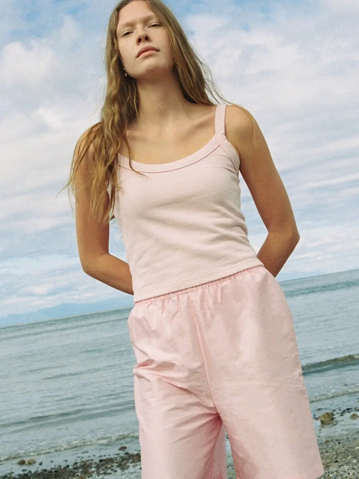 A model stands outdoors by the ocean wearing a casual Merge Goods Hemp Breeze Tank Top in Pink Cream with wide, matching light pink athletic shorts under a cloudy sky | Twentyseven Toronto