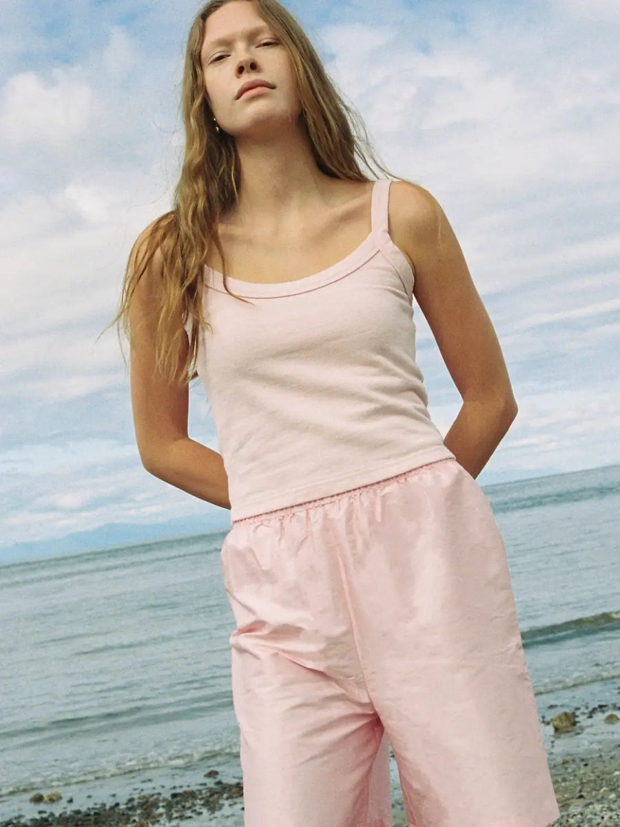 A model stands outdoors by the ocean wearing a casual Merge Goods Hemp Breeze Tank Top in Pink Cream with wide, matching light pink athletic shorts under a cloudy sky | Twentyseven Toronto