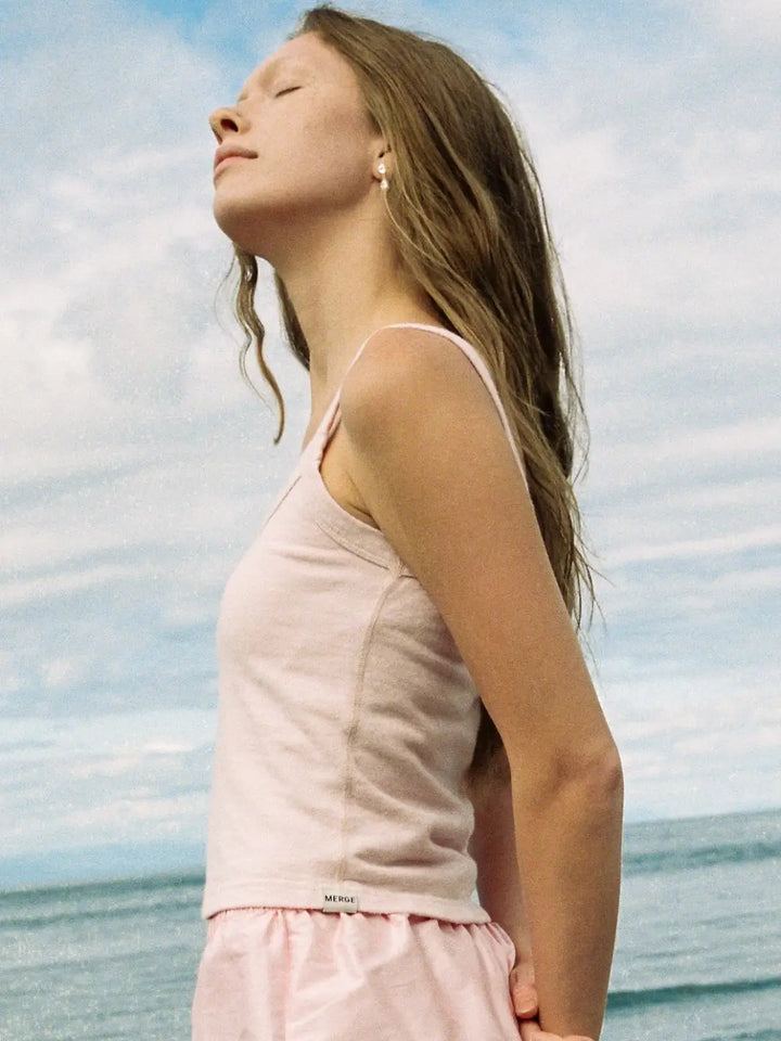 A side-view of a woman looking upwards against a coastal background while wearing a fitted, light pink Merge Goods Hemp Breeze Tank Top in Pink Cream | Twentyseven Toronto