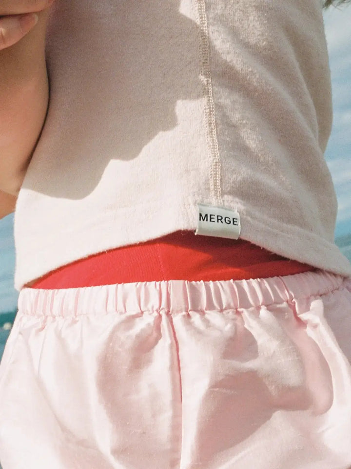 A close-up focuses on the small white brand label stitched onto the bottom hem of the light pink Merge Goods Hemp Breeze Tank Top in Pink Cream | Twentyseven Toronto