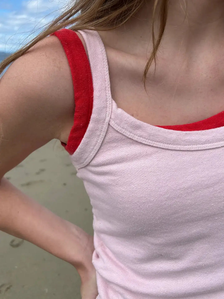 A close-up shot shows the neckline of a light pink Merge Goods Hemp Breeze Tank Top in Pink Cream worn over a vibrant red top on a sandy beach | Twentyseven Toronto