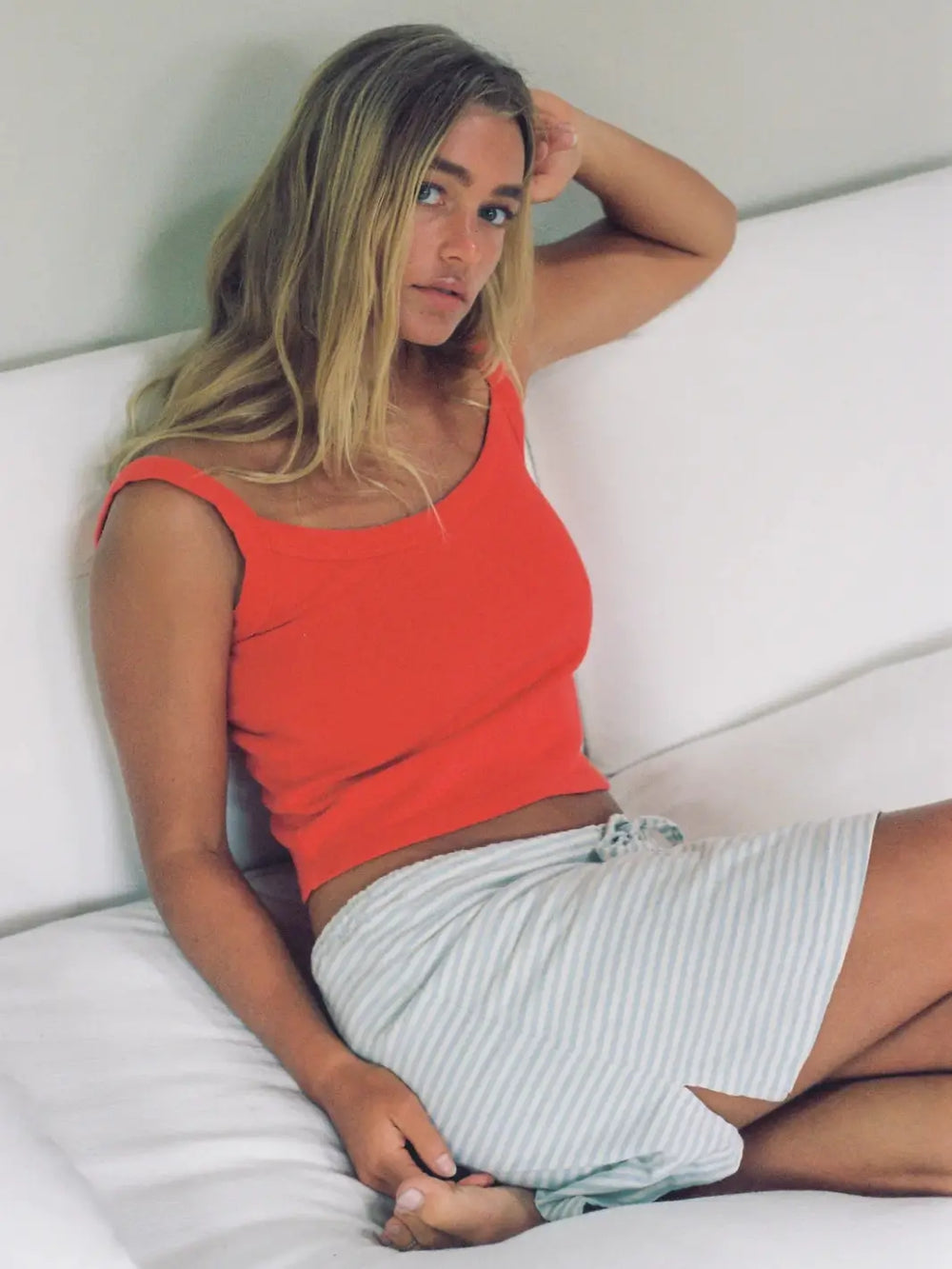A woman reclines on a white sofa wearing a bright Merge Goods Hemp Breeze Tank Top in Poppy Red paired with blue and white striped shorts | Twentyseven Toronto