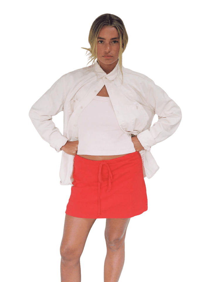 A model wearing the bright red Merge Goods Hemp Lazy Skort in Poppy Red under a white top and an open white button-down jacket | Twentyseven Toronto