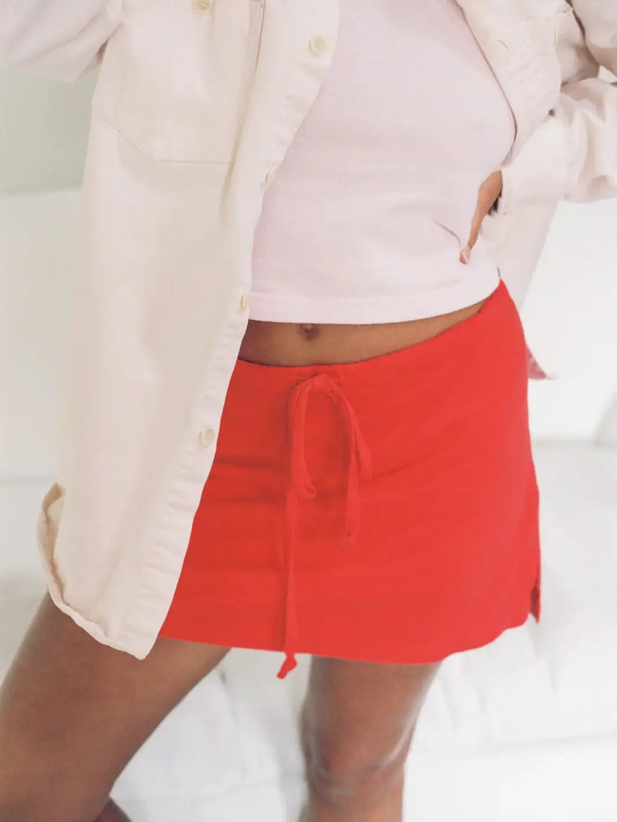 A close-up view displays a person wearing the bright red Merge Goods Hemp Lazy Skort in Poppy Red with a drawstring waist, styled under a white tank top and an unbuttoned cream shirt | Twentyseven Toronto