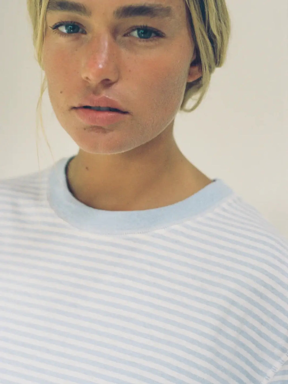 A close-up shot shows a woman with blonde hair wearing the crewneck Merge Goods Hemp Rover Tee in Daydream Stripes featuring light blue and white horizontal pinstripes | Twentyseven Toronto