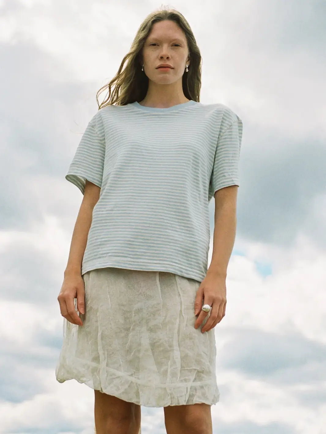A person poses outdoors under a cloudy sky wearing a relaxed-fit Merge Goods Hemp Rover Tee in Daydream Stripes paired with a textured white midi skirt | Twentyseven Toronto