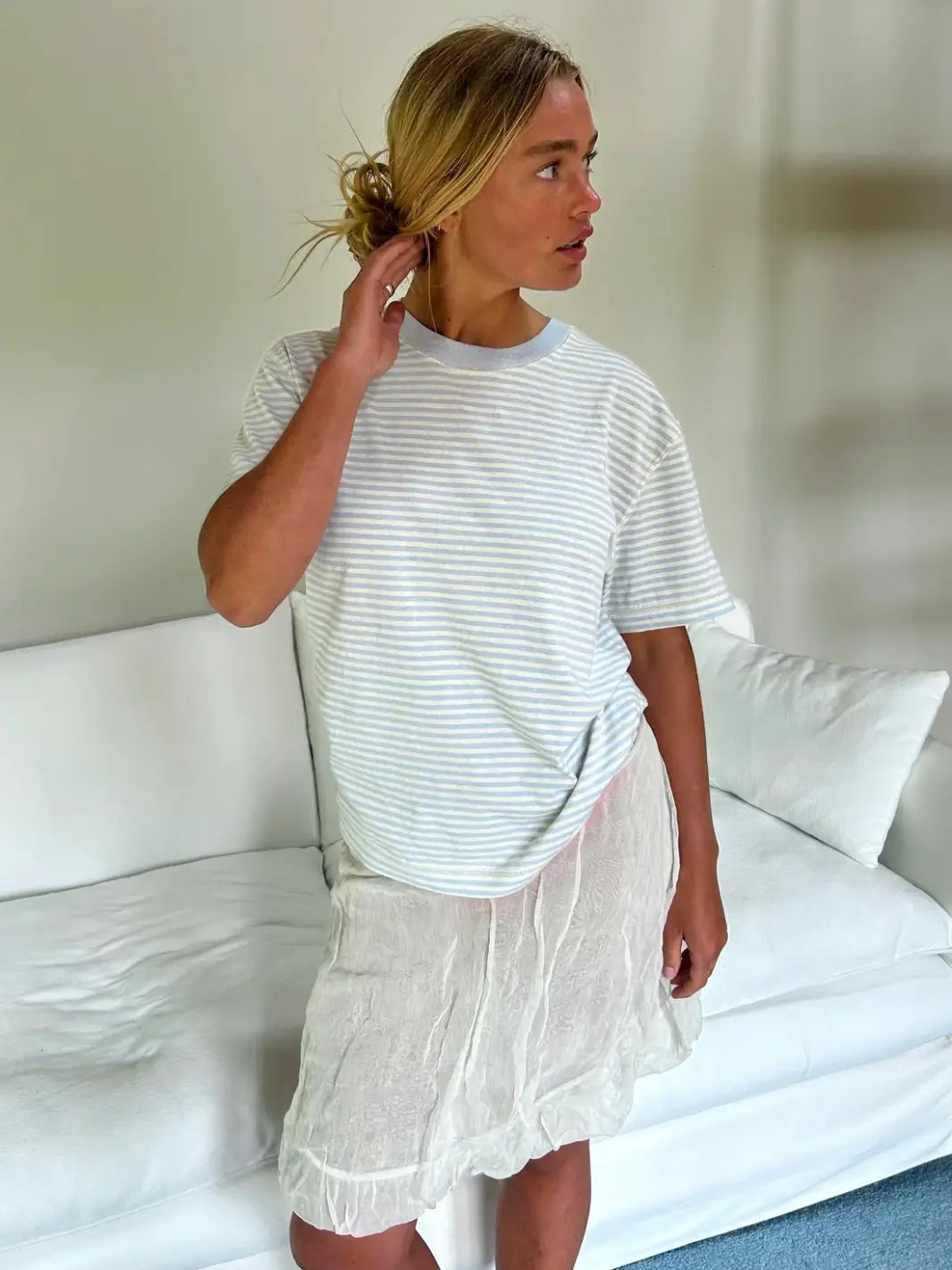 A person stands in front of a white sofa wearing a loose-fitting Merge Goods Hemp Rover Tee in Daydream Stripes with a light blue ribbed collar and a semi-sheer white skirt | Twentyseven Toronto