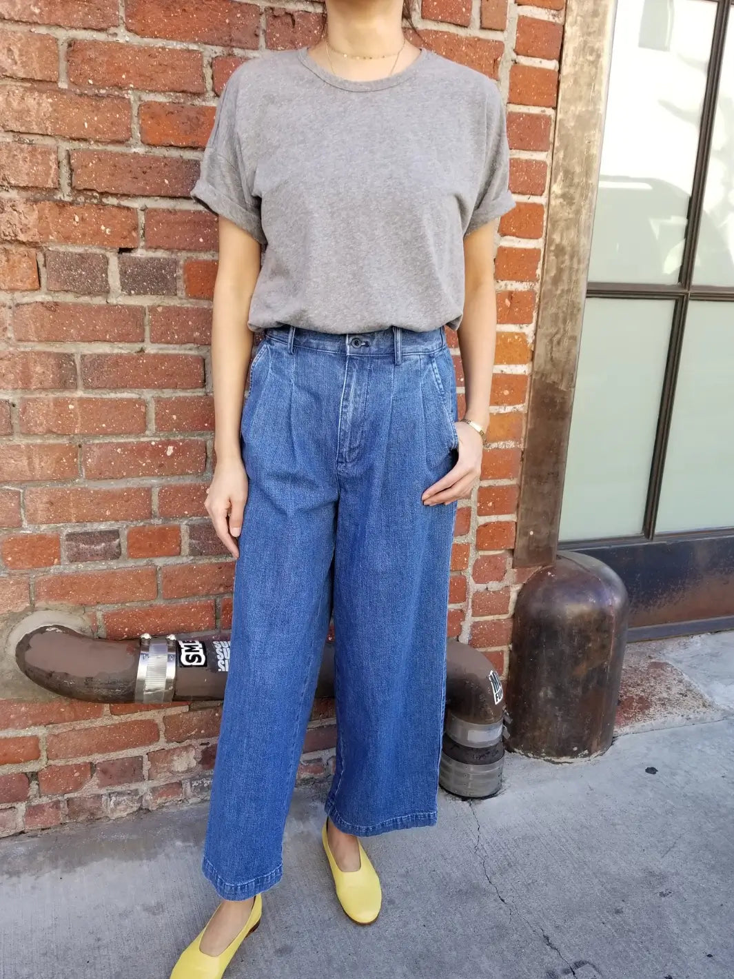Person wearing a grey Her Tee shirt in 'HT Grey' from Le Bon Shoppe and blue jeans standing outdoors against a brick background | Twentyseven Toronto
