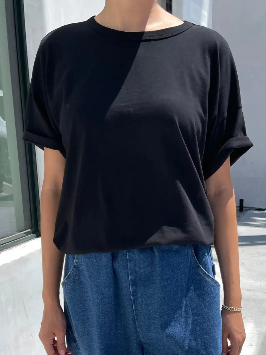 A person stands outdoors wearing the crewneck Le Bon Shoppe Her Tee - True Black featuring cuffed short sleeves and a relaxed fit paired with blue denim jeans | Twentyseven Toronto