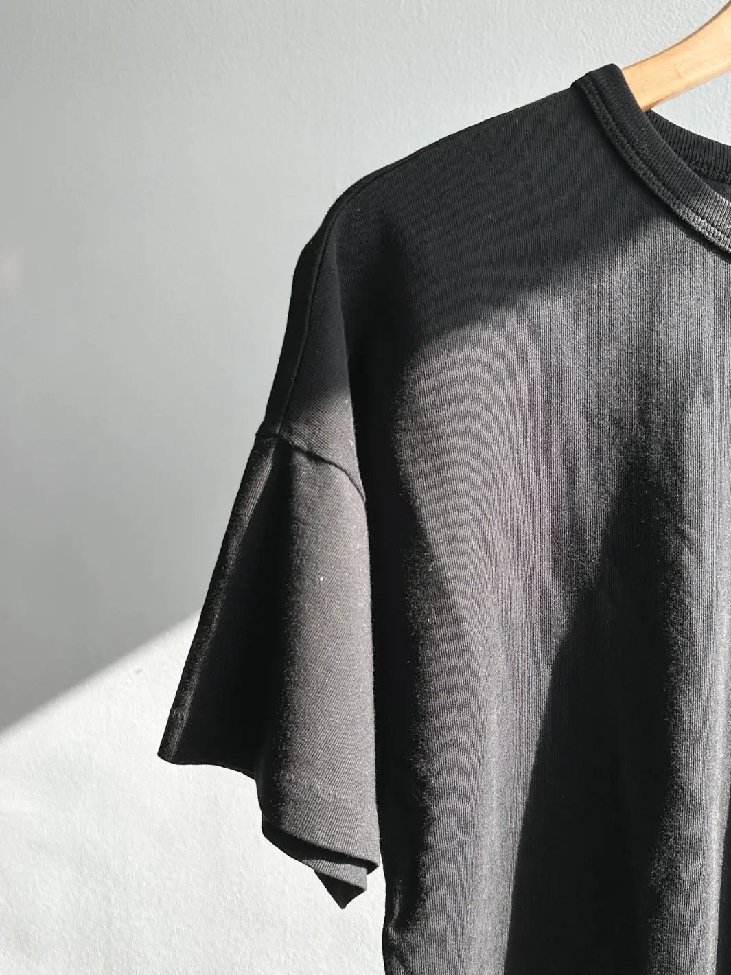 A close-up, angled shot showcases the fine fabric texture and drop-shoulder sleeve construction of the Le Bon Shoppe Her Tee - True Black hanging on a wooden hanger against a white wall | Twentyseven Toronto