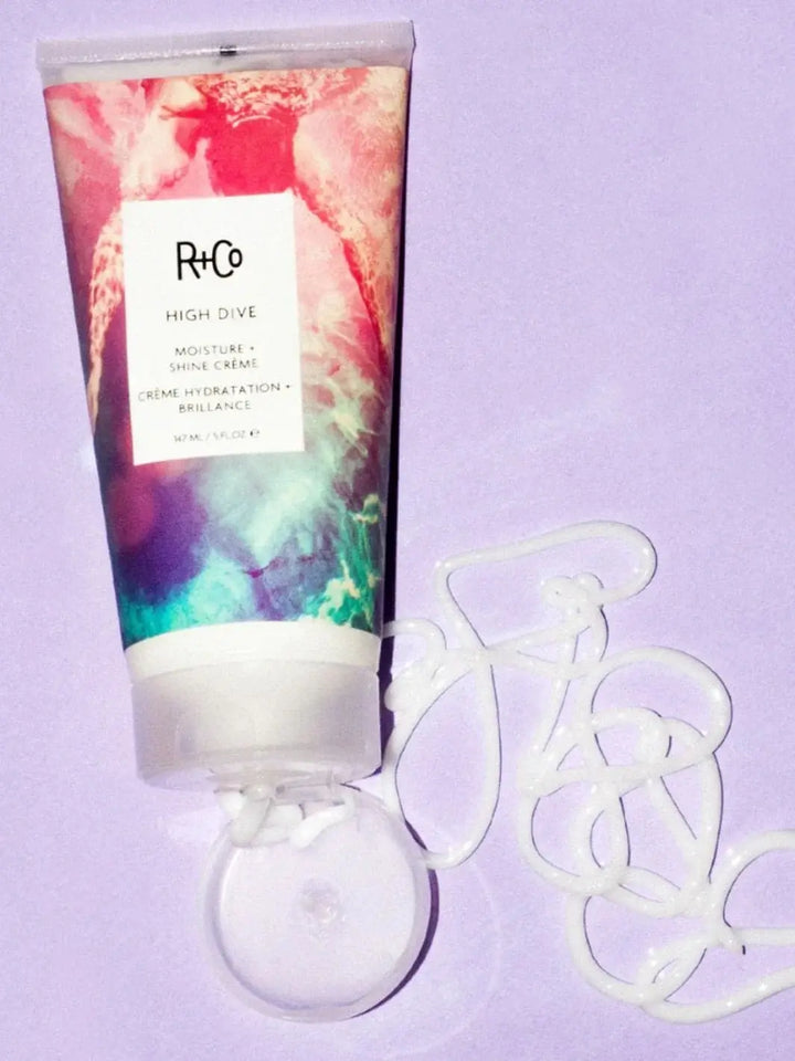 A squeeze tube of R+Co HIGH DIVE Moisture & Shine Crème lays next to a decorative swirl of the white hair cream on a light purple surface | Twentyseven Toronto