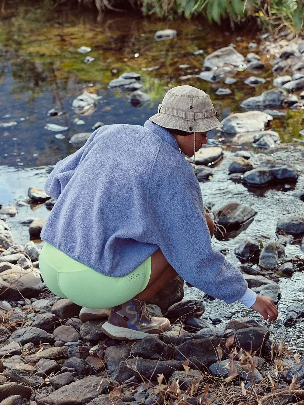 A person crouching by a rocky stream wearing a periwinkle blue Free People Hit The Slopes Colorblock Jacket - Trailblazer Combo, lime green shorts, and a bucket hat | Twentyseven Toronto