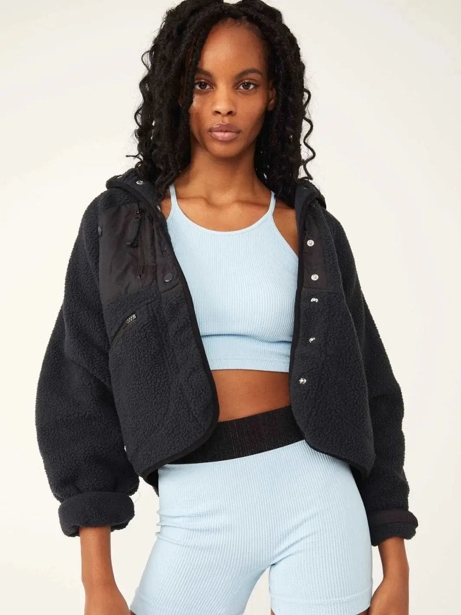 A model wears an open, button-front Free People Hit The Slopes Fleece Jacket - Black layered over a light blue athletic crop top and shorts set | Twentyseven Toronto