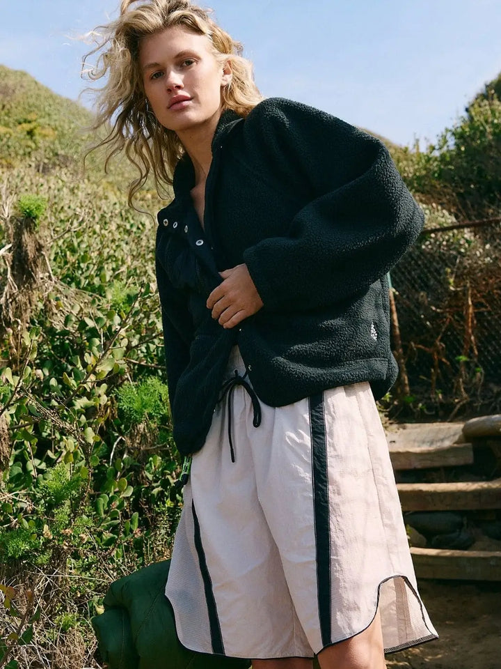 A model stands outdoors on a path wearing the Free People Hit The Slopes Fleece Jacket - Black styled with light-colored athletic shorts | Twentyseven Toronto