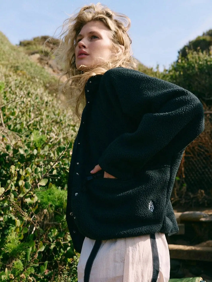 A rear view highlights the high collar and drop-shoulder sleeve design of the Free People Hit The Slopes Fleece Jacket - Black as the model holds a rolled green mat | Twentyseven Toronto