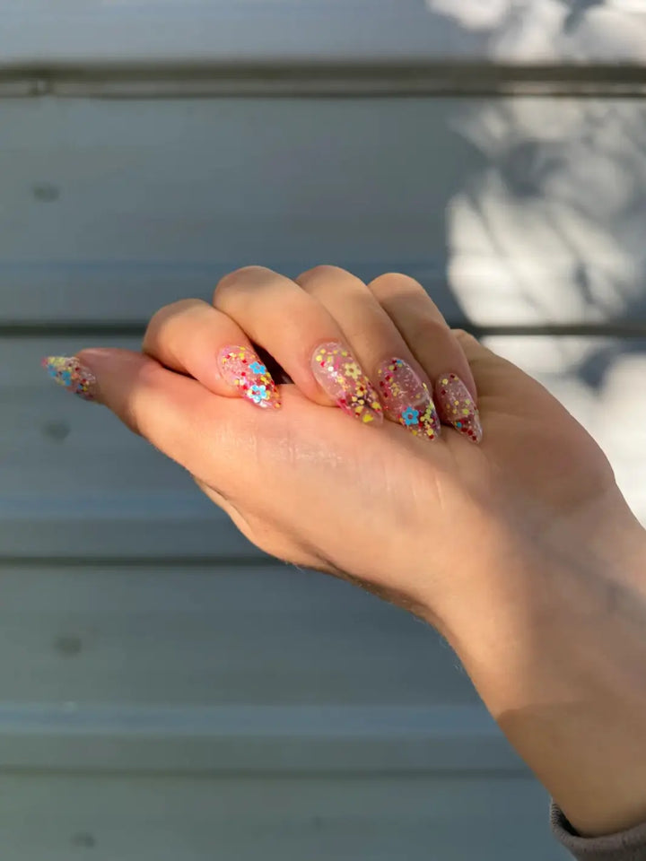 Close-up of a hand with glittery pastel pink, blue and yellow 'Hot Pants' nail polish by Death Valley Nails against a blurred natural background | Twentyseven Toronto