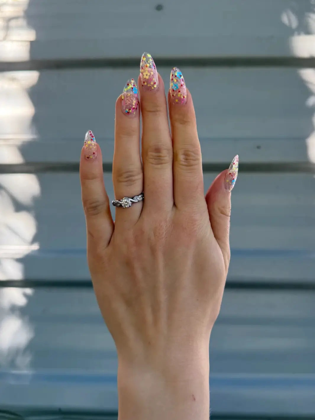 Close-up of a hand with glittery pastel pink, blue and yellow 'Hot Pants' nail polish by Death Valley Nails against a blurred natural background | Twentyseven Toronto