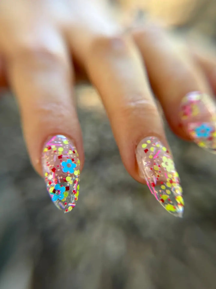 Close-up of a hand with glittery pastel pink, blue and yellow 'Hot Pants' nail polish by Death Valley Nails against a blurred natural background | Twentyseven Toronto