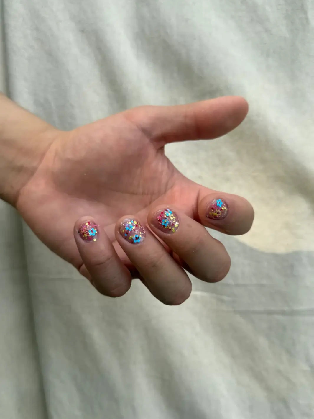 Close-up of a hand with glittery pastel pink, blue and yellow 'Hot Pants' nail polish by Death Valley Nails against a blurred natural background | Twentyseven Toronto