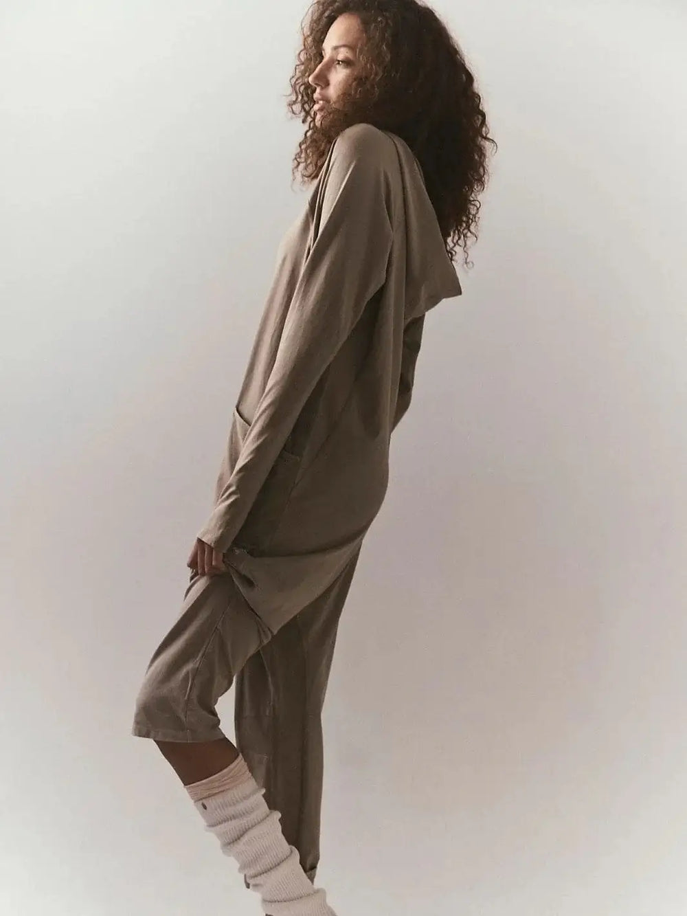 A side-angle view captures the comfortable, long-sleeve silhouette of the Free People Hot Shot Long Sleeve Onesie - Sage Stone styled with slouchy white leg warmers | Twentyseven Toronto