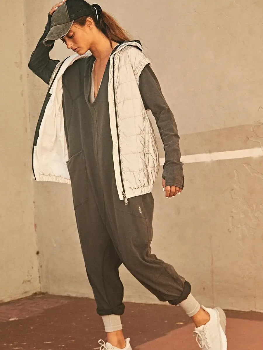 A model poses outside wearing a washed black Free People Hot Shot Long Sleeve Onesie - Black layered with a light-colored quilted vest, baseball cap, and sneakers | Twentyseven Toronto
