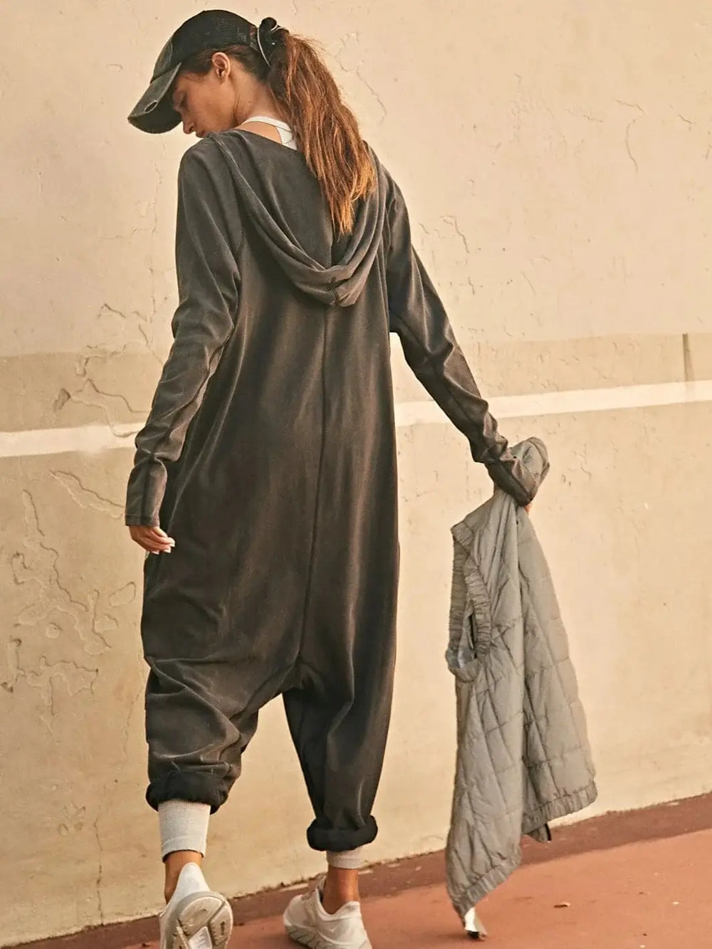 A view from behind shows the hood, relaxed fit, and dropped-crotch design of the Free People Hot Shot Long Sleeve Onesie - Black as the model walks away holding a vest | Twentyseven Toronto