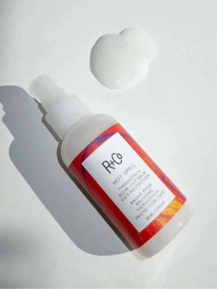A bottle of R+Co HOT SPELL ThermoTech Blow Out Balm 450° F Protection lies diagonally on a white surface next to a dollop of the smooth, white cream | Twentyseven Toronto