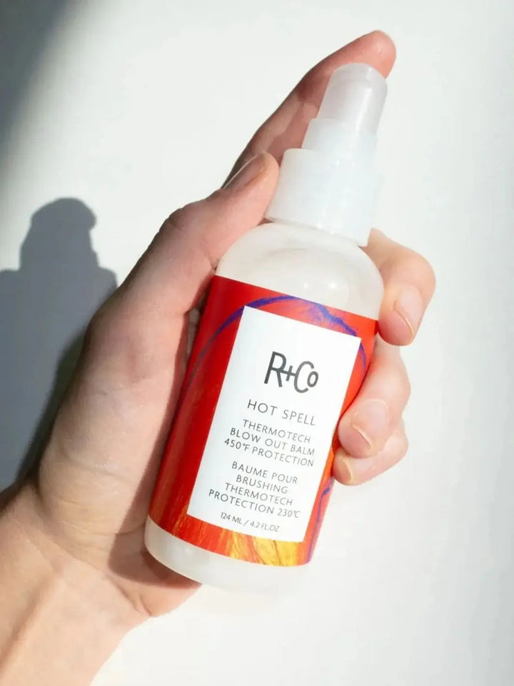 A hand holds up a bottle of R+Co HOT SPELL ThermoTech Blow Out Balm 450° F Protection against a clean white background | Twentyseven Toronto