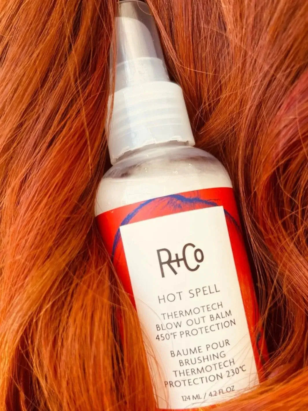 A close-up shot shows a bottle of R+Co HOT SPELL ThermoTech Blow Out Balm 450° F Protection nestled amidst vibrant, sleek copper-red hair strands | Twentyseven Toronto