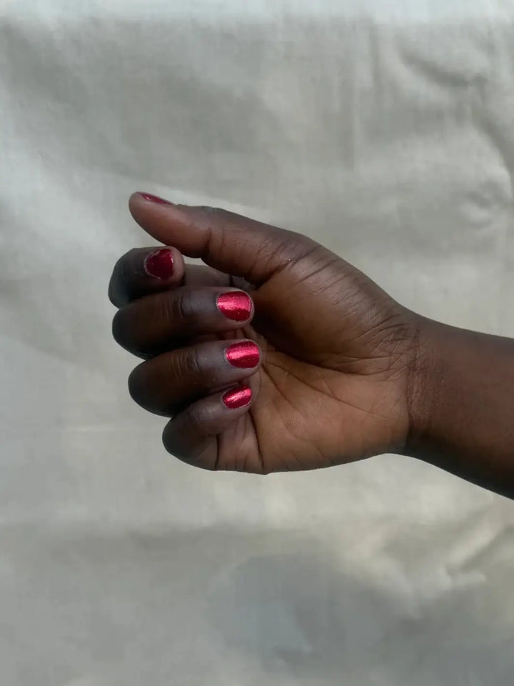 A close-up view of a hand with a deep skin tone shows short nails painted in the vibrant, metallic red of Death Valley Nails Hot Tamale Nail Polish | Twentyseven Toronto