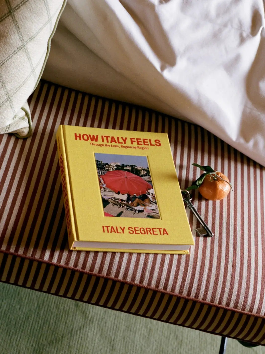 A lifestyle photo of the book How Italy Feels: Through the Lens, Region by Region by Italy Segreta resting on a brown-and-white striped cushion next to a key and clementine | Twentyseven Toronto