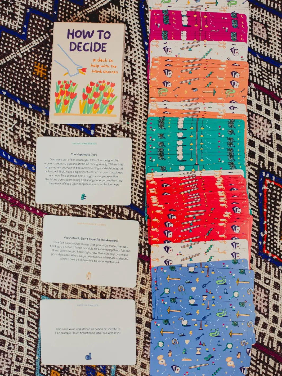 The How To Decide Deck from People I've Loved box sits next to a vertical stack of fanned card backs and three revealed text cards on a woven, geometric rug | Twentyseven Toronto