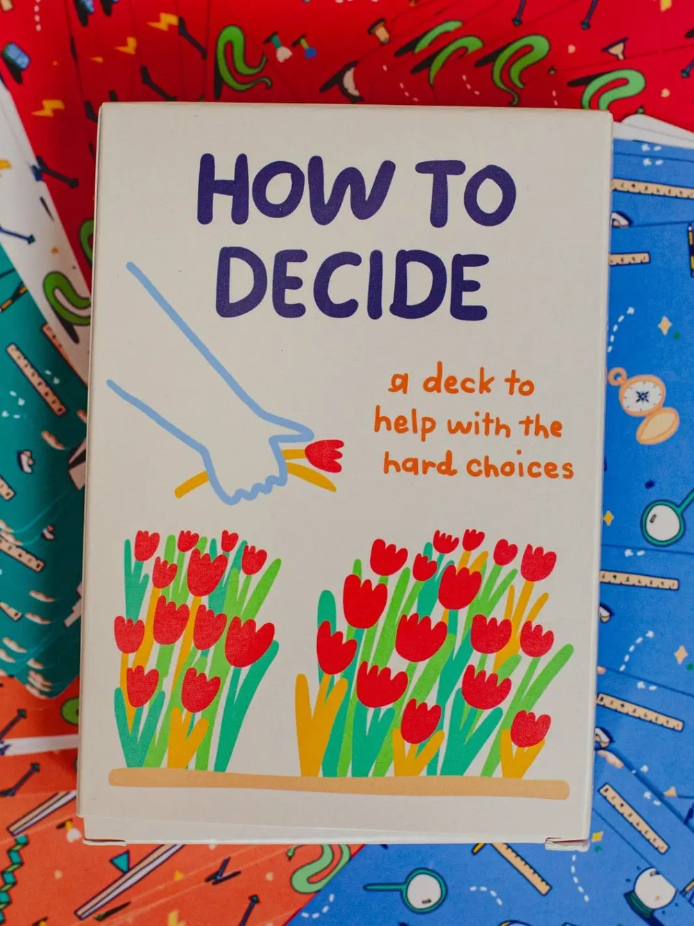The front box of the How To Decide Deck from People I've Loved is displayed face-up against a vibrant background of colourful, patterned playing cards | Twentyseven Toronto