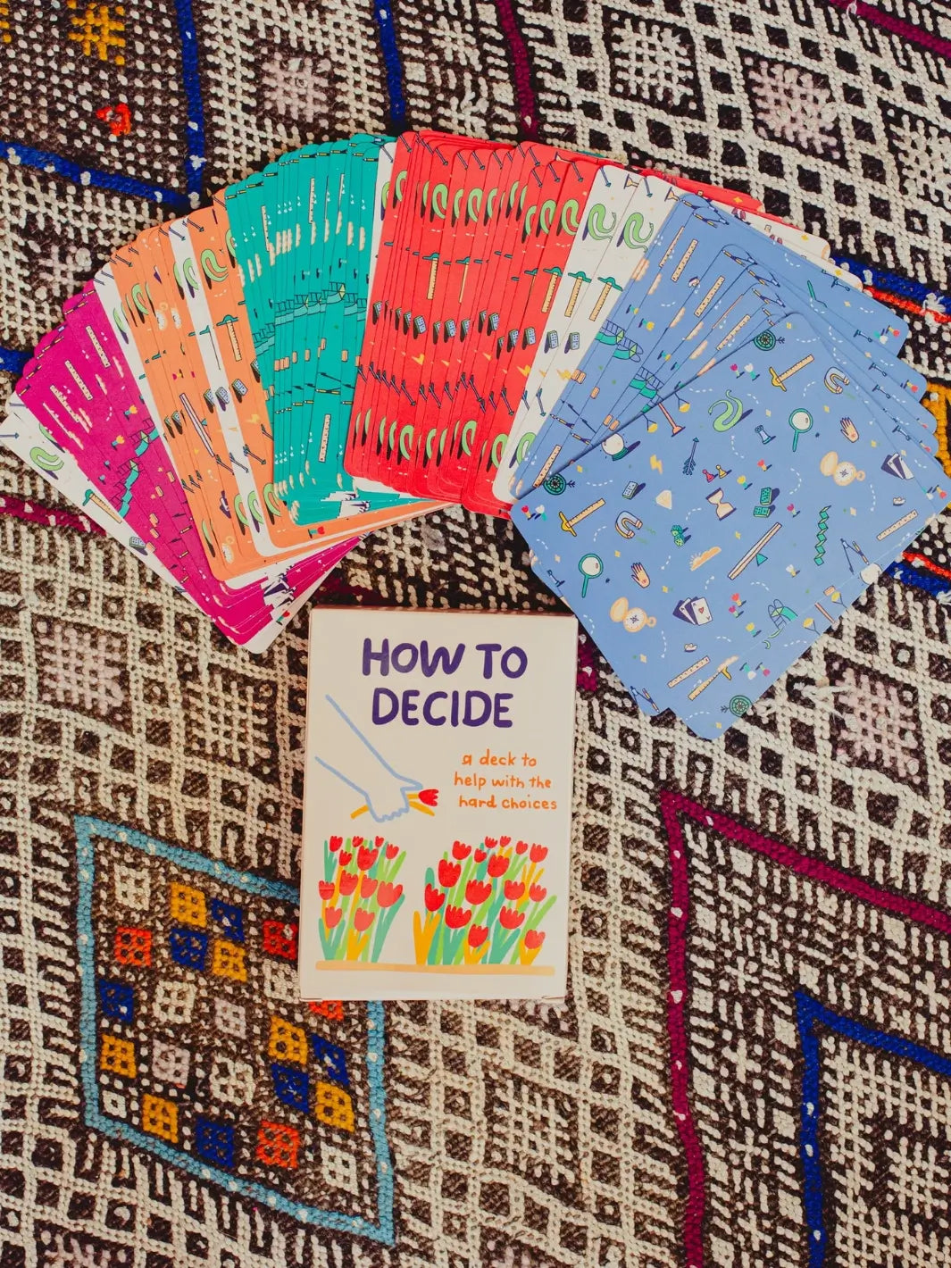 The card box for the How To Decide Deck from People I've Loved sits on a patterned rug beneath a large, fanned-out arrangement of the colourful patterned card backs | Twentyseven Toronto