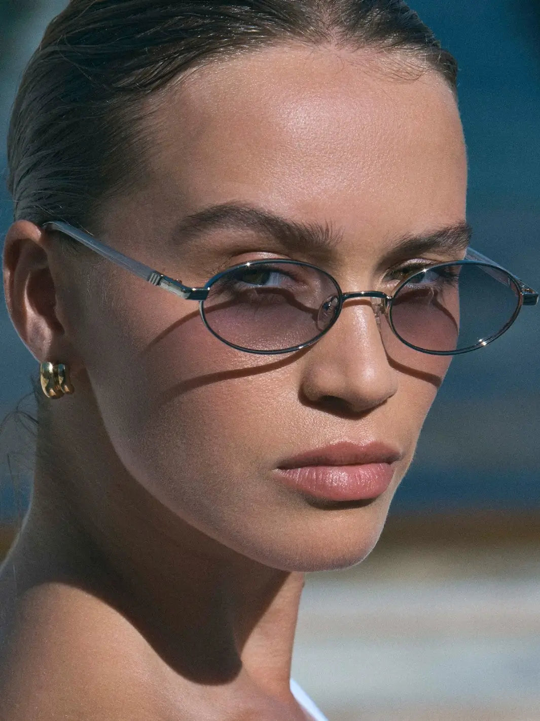 An outdoor, close-up shot of a woman wearing silver, oval-framed Le Specs Icicle - Silver Smoke Grad sunglasses | Twentyseven Toronto