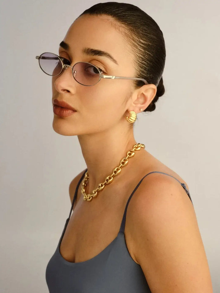 A woman wearing the oval Le Specs Icicle - Silver Smoke Grad sunglasses paired with a chunky gold chain necklace | Twentyseven Toronto