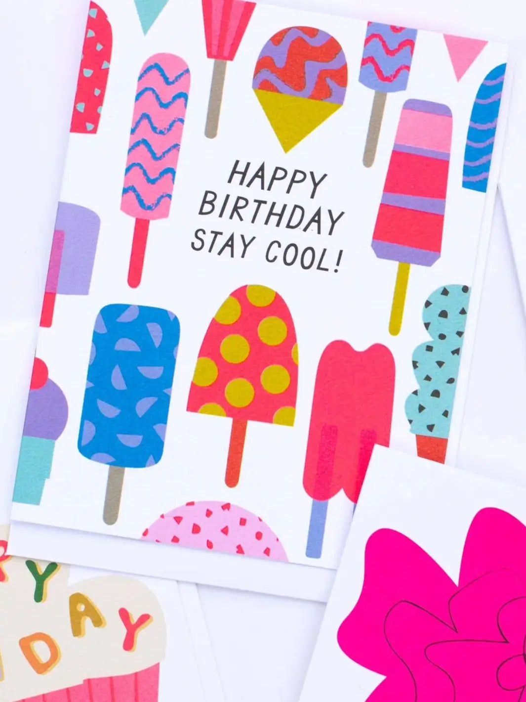 An angled, close-up shot shows the Banquet Workshop Icy Treats Happy Birthday Stay Cool Card displayed on a white surface overlapping other colorful illustrated greeting cards | Twentyseven